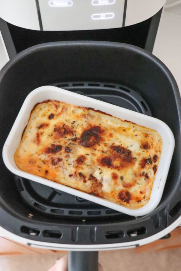 A white rectangular dish with a single serving of zucchini lasagna, topped with melted cheese and herbs, is placed inside an air fryer basket. The air fryer display is visible in the background.