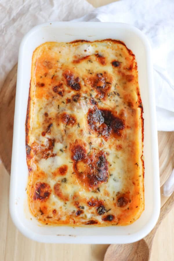 A single serving of zucchini lasagna in a white rectangular dish, with melted and slightly browned cheese on top, is set on a wooden surface. A wooden spoon and a white cloth are nearby.