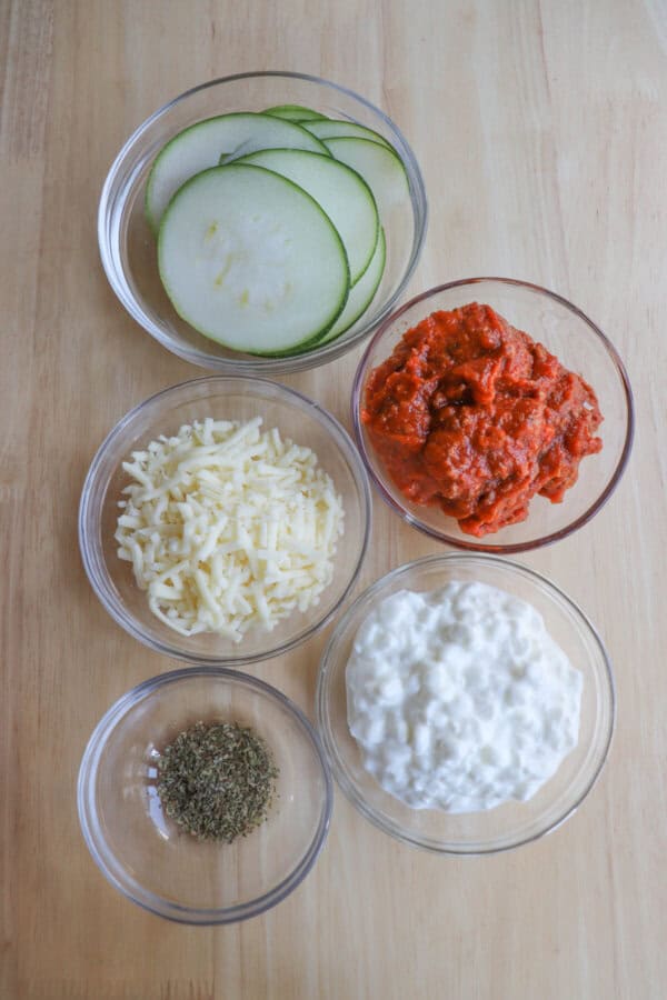 A wooden surface showcases a delightful single serving zucchini lasagna setup, featuring bowls filled with sliced zucchini, tomato sauce, shredded cheese, cottage cheese, and dried herbs.