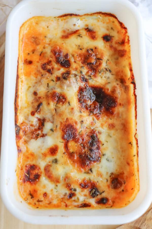 A baked single serving of zucchini lasagna rests in a white rectangular dish. The top layer is bubbly and golden brown, with melted cheese and herbs visible, creating a slightly crispy appearance. This enticing dish sits on a wooden surface, ready to be enjoyed.