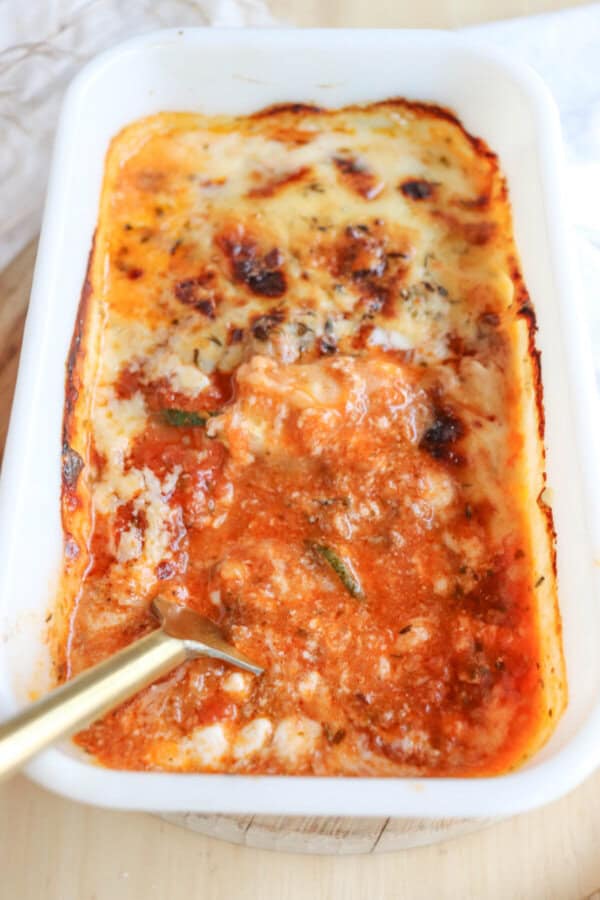 A baking dish with a portion of single serving zucchini lasagna removed reveals layers of sauce, cheese, and pasta. The top is browned and bubbly. A gold-colored spoon rests in the dish, ready to serve another delicious bite.