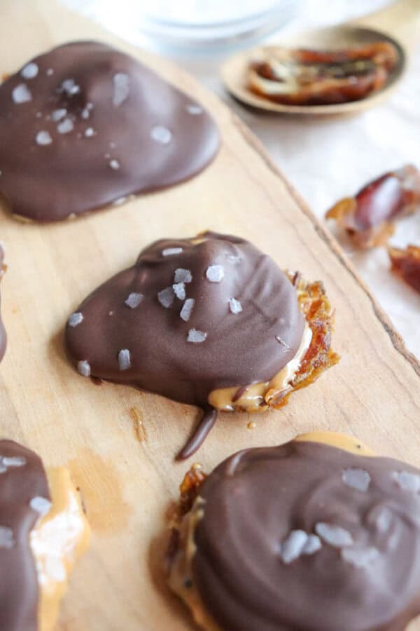Chocolate-covered treats with a caramel center are sprinkled with sea salt on a wooden board. They are accompanied by natural dates, one partially visible in the background.