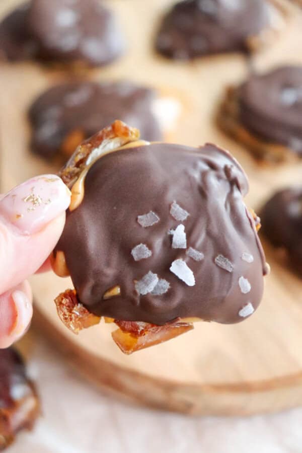 A hand holding a chocolate-covered date treat topped with flaky sea salt. The treat has a glossy coating and is placed above a wooden board with more similar treats in the background.