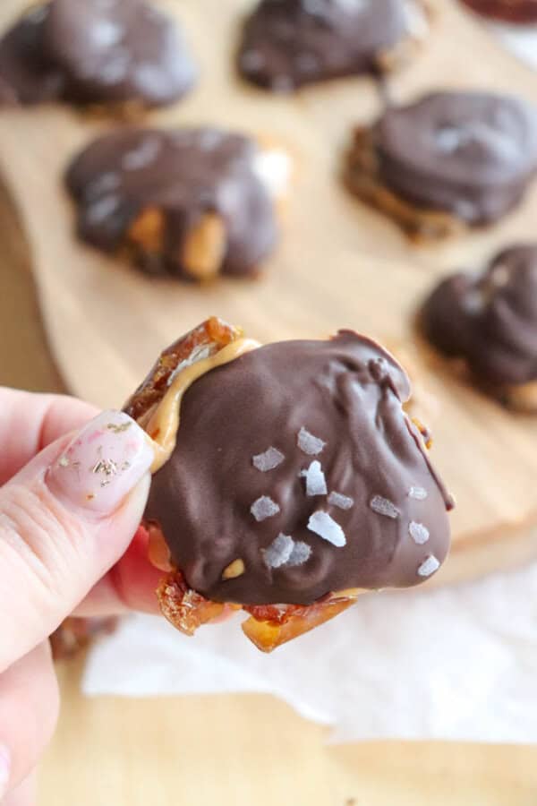 A hand holds a chocolate-covered treat topped with sea salt flakes. Behind it, more similar treats are placed on a wooden board. The treat appears to have a chewy base, possibly made of caramel or nougat.
