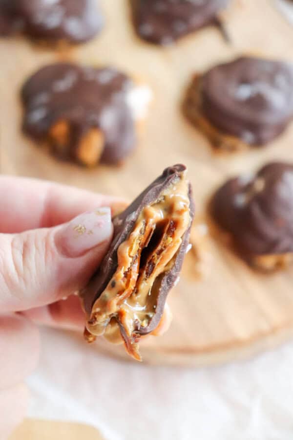 A close-up of a hand holding a chocolate-covered treat with a visible cross-section showing layers of date and peanut butter. Other similar treats are blurred in the background on a wooden surface.