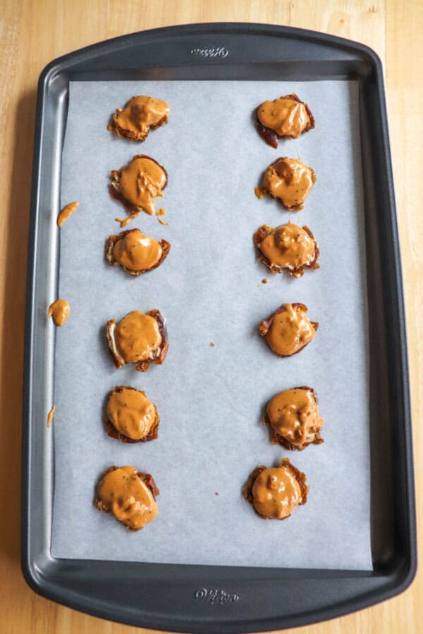 A baking sheet lined with parchment paper holds three rows of smashed dates, topped with dollops of peanut butter. The dates are arranged neatly in a grid pattern on the sheet.