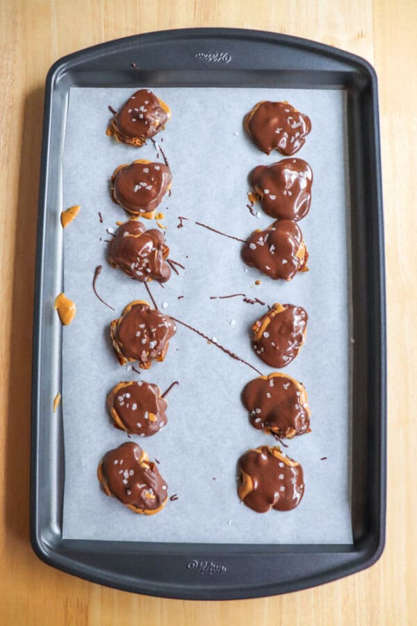 A baking tray with parchment paper holds two rows of date bites drizzled with chocolate. The treats have a sprinkle of sea salt on top, placed on a wooden surface.