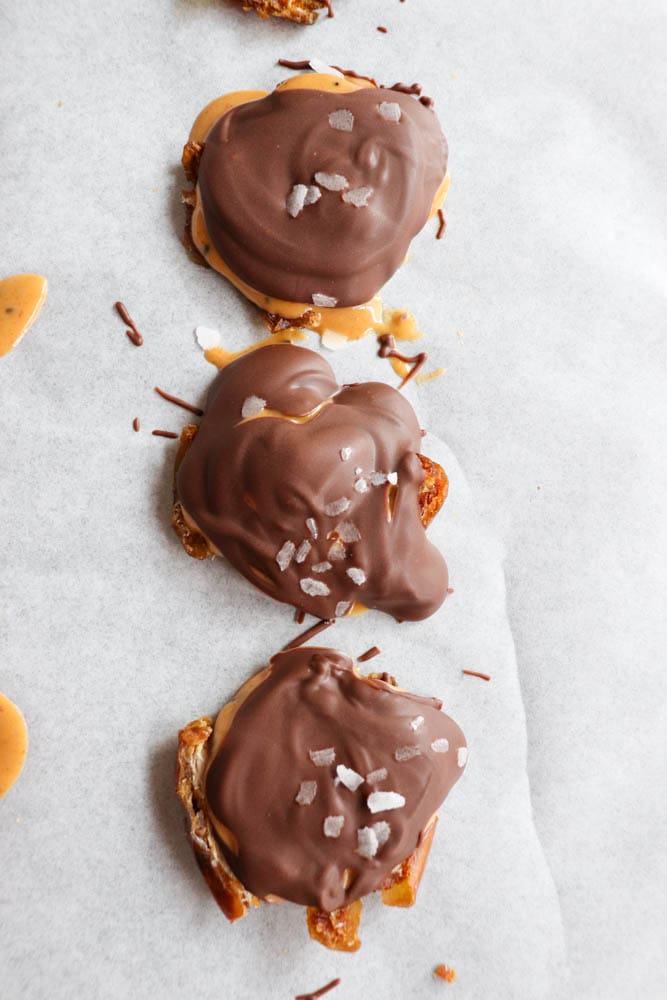 Three chocolate-covered treats are lined up on parchment paper. Each treat has a smooth chocolate coating sprinkled with a few flakes of sea salt. The shapes are irregular, and a bit of the filling is visible on the edges.