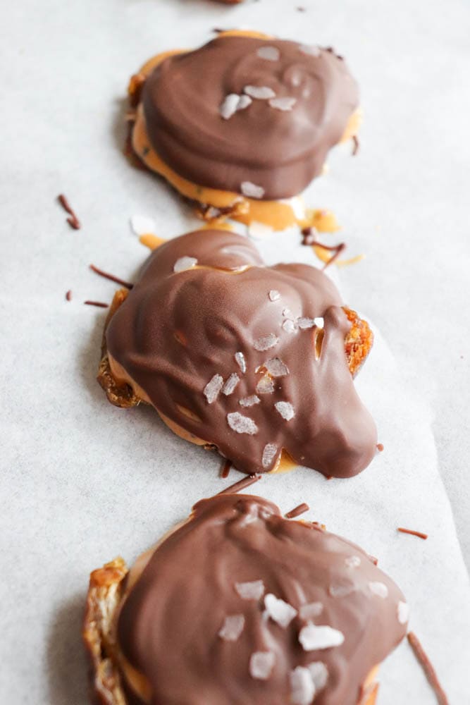 Four chocolate-covered treats are arranged in a line on parchment paper. Each treat is topped with a sprinkle of coarse sea salt, creating a contrast against the smooth chocolate coating. The treats appear to have a caramel layer beneath the chocolate.