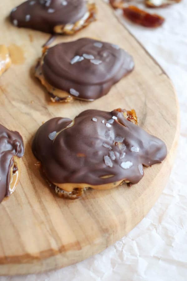 Chocolate-covered dates topped with sea salt flakes are placed on a wooden board. The treats are glossy and appear to have a chewy texture underneath the chocolate coating.