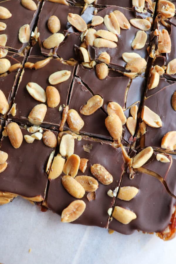 Close-up of chocolate Snickers squares topped with whole peanuts. The treats are cut into sections, revealing a glossy chocolate layer sprinkled with salted peanuts. They rest on a sheet of baking paper.