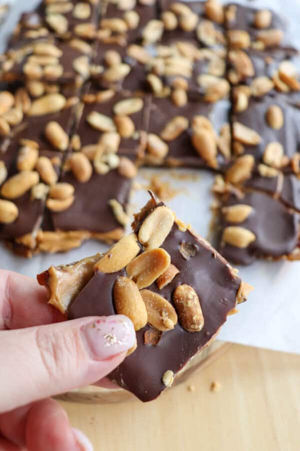 A hand holding a square piece of chocolate-covered date bark topped with peanuts. In the background, more pieces of the toffee are placed on parchment paper. The persons nails are painted with a light color and delicate floral design.