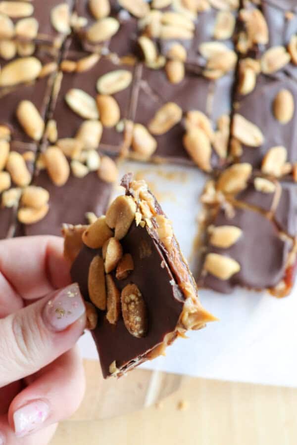 A hand holding a square piece of chocolate bark topped with peanuts. In the background, more pieces of the chocolate bark with scattered peanuts are visible on parchment paper.
