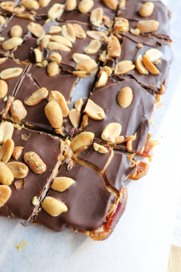A slab of chocolate-covered dates cut into square pieces, topped with whole peanuts. The dessert appears to be resting on parchment paper, highlighting the rich chocolate layer and crunchy peanuts.