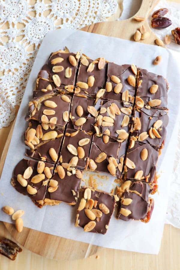 Square pieces of chocolate-covered bars topped with whole peanuts are arranged on parchment paper on a wooden board. Some crumbs and loose peanuts are scattered around. A lace doily is partially visible on the table.