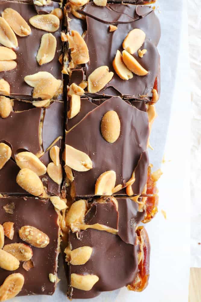 Close-up of chocolate-topped bars, sliced into squares. The bars are topped with whole peanuts and a smooth layer of chocolate, revealing a date filling beneath. They rest on parchment paper on a light surface.