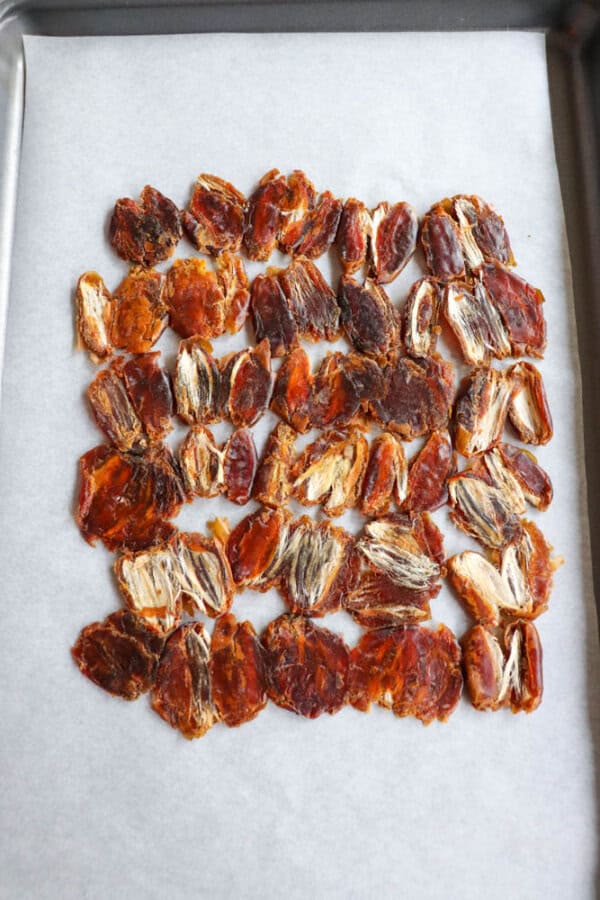 A baking sheet lined with parchment paper holds several rows of halved, pitted dates. The dates vary in color from deep brown to a lighter orange-brown, showcasing their natural texture and fibrous interiors.