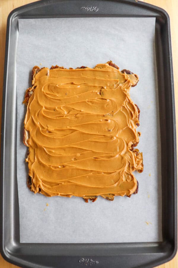 A rectangular sheet of parchment paper on a baking tray, topped with a layer of smooth, swirled caramel or peanut butter spread evenly. The spread covers a date base underneath.