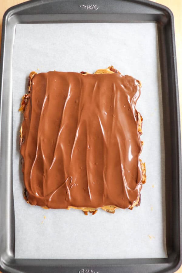 A baking sheet holds a piece of parchment paper with a rectangular dessert topped with a smooth layer of melted chocolate.