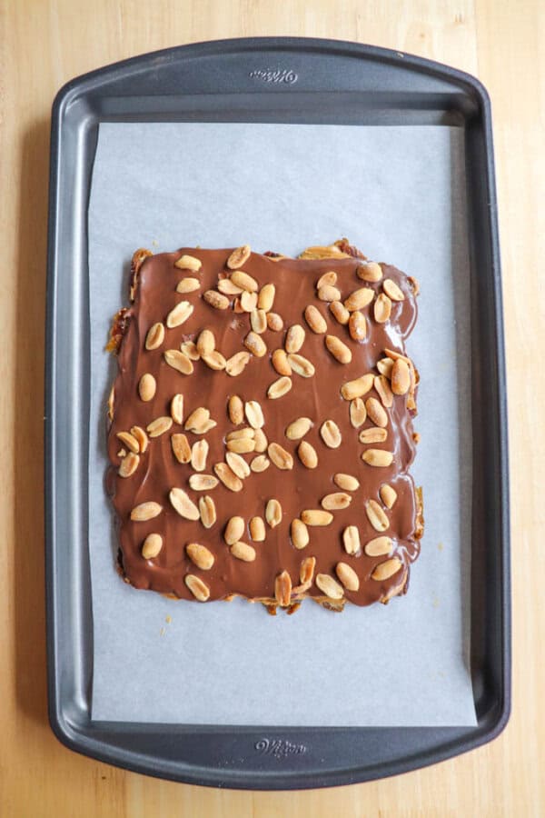 Baking tray with a square layer of chocolate-covered dessert topped with scattered peanuts on parchment paper.