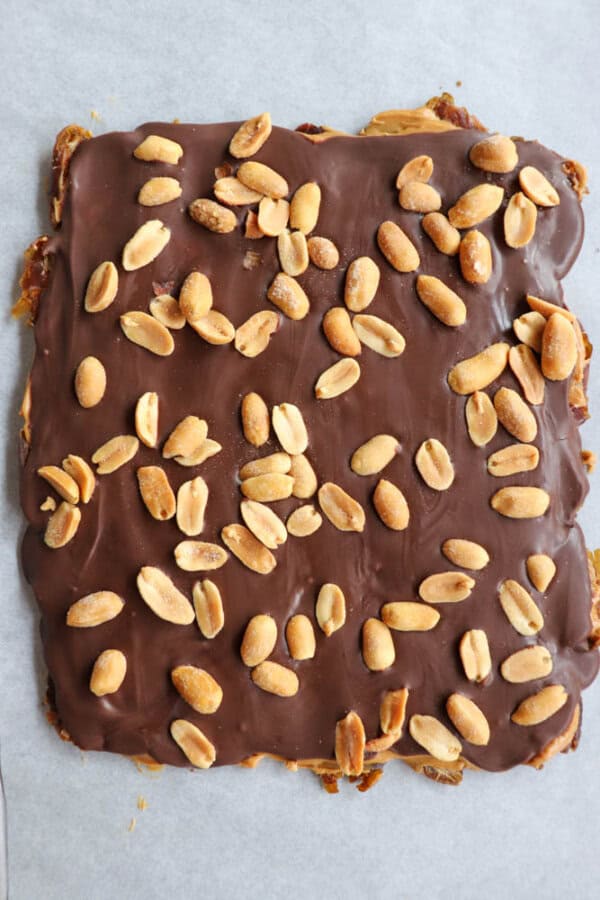 A square dessert topped with smooth chocolate and sprinkled with roasted peanuts, resting on a parchment paper background.