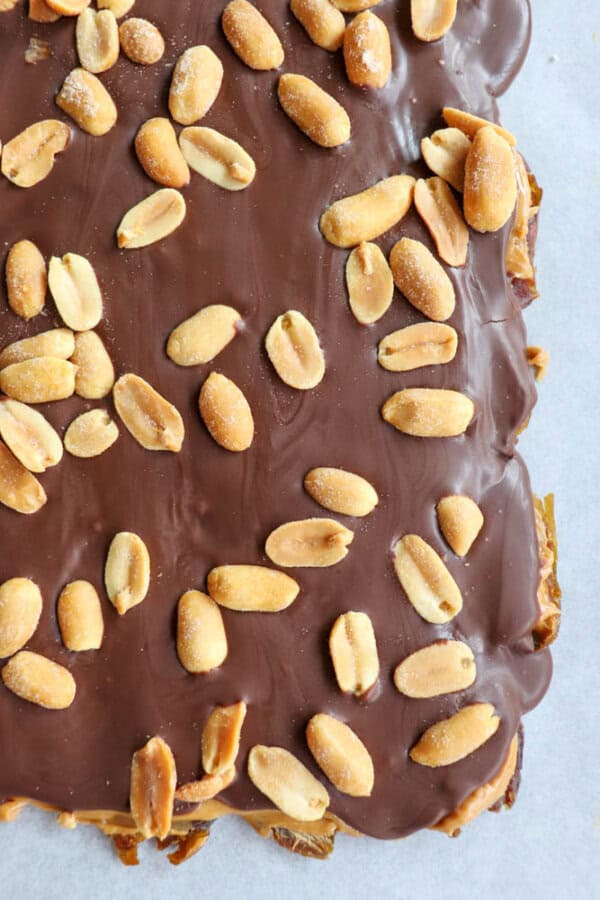 Close-up of a chocolate-covered dessert topped with scattered peanuts on a parchment paper surface. The smooth chocolate layer is generously sprinkled with whole peanuts, creating a sweet and nutty treat.