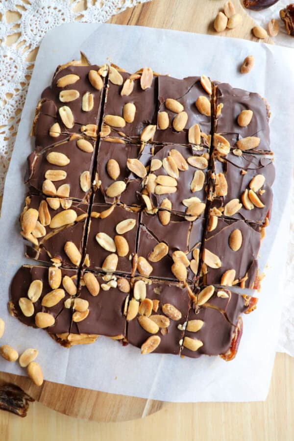A batch of chocolate-covered snickers date bark squares topped with whole peanuts, cut into 16 pieces, rests on a piece of parchment paper on a wooden cutting board. Some scattered peanuts are visible around the board.