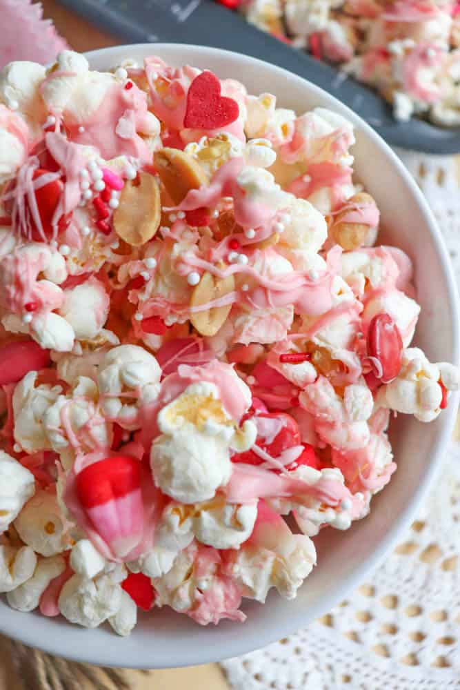 A bowl filled with popcorn coated in pink and white drizzle, mixed with red and heart-shaped candies, and small sprinkles. The colorful mix is placed on a textured white surface.
