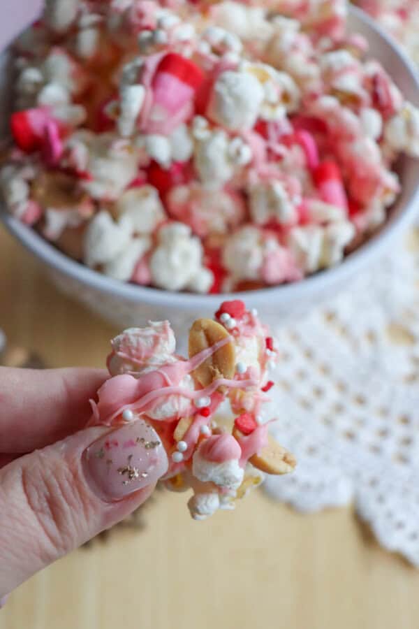 A hand holding a piece of popcorn snack mix coated in pink, white, and red sprinkles with small candy pieces. A bowl holds more of the colorful mix in the background, set on a light wooden table with a lace doily.