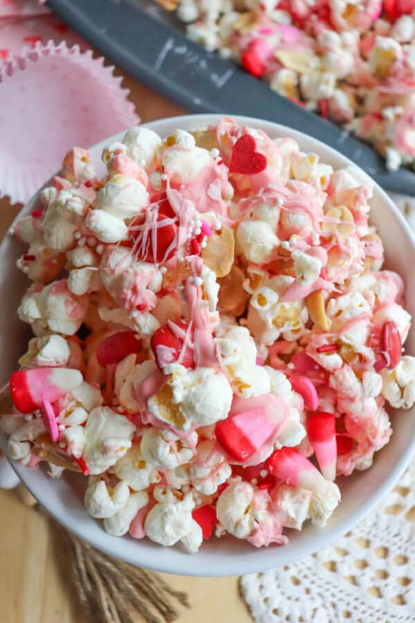 A bowl of popcorn mixed with heart-shaped red and pink candies, drizzled with pink and white icing. The vibrant colors create a festive and sweet look, perfect for a celebration or themed event.