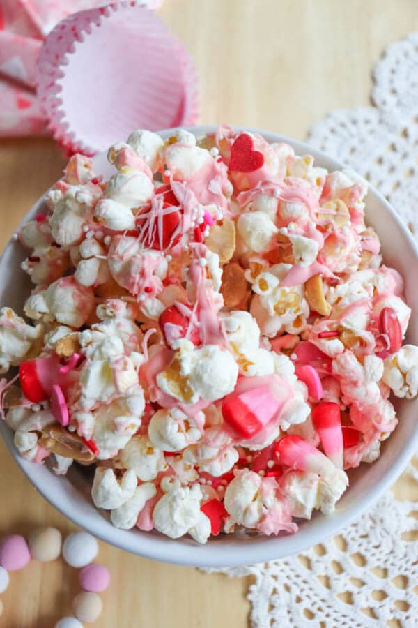 A bowl of popcorn mixed with pink and red candies, drizzled with pink chocolate and topped with heart-shaped sprinkles. The bowl is placed on a wooden table with doilies, cupcake liners, and small round candies nearby.