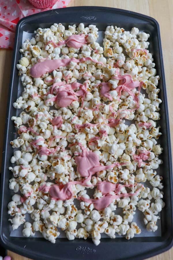 A tray of popcorn drizzled with pink icing, spread out on parchment paper. The tray is on a wooden surface, and there are pink and white polka dot cupcake liners in the background.