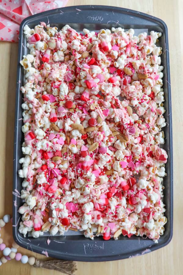 A baking tray filled with a colorful mix of popcorn, mini marshmallows, and various toppings. The mixture includes pink and red sprinkles, chocolate drizzle, and small biscuit pieces, all on a light wooden surface.