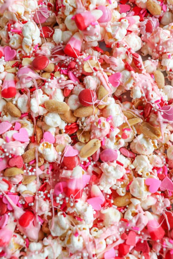 A vibrant mix of popcorn, pink and red candies, peanuts, and heart-shaped sprinkles, all drizzled with pink and white chocolate, creating a festive and colorful snack blend.