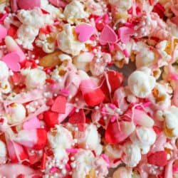 A colorful mix of popcorn covered in pink candy coating, with heart-shaped and round sprinkles. Various shades of pink and red candies are scattered throughout, creating a festive and sweet appearance.