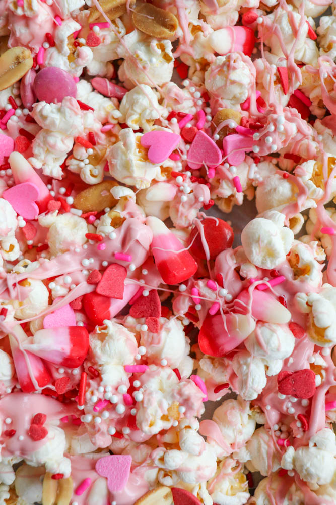 A colorful mix of popcorn covered in pink candy coating, with heart-shaped and round sprinkles. Various shades of pink and red candies are scattered throughout, creating a festive and sweet appearance.