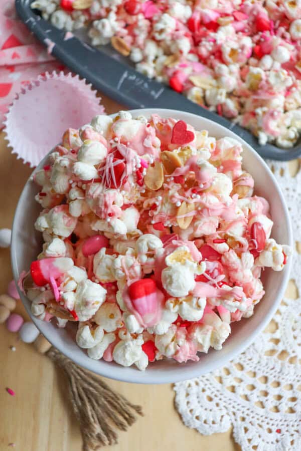 A bowl of popcorn drizzled with pink and red candy melts, topped with heart-shaped sprinkles and candy pieces. Its placed on a wooden surface with doily accents and a baking tray of similar popcorn in the background.