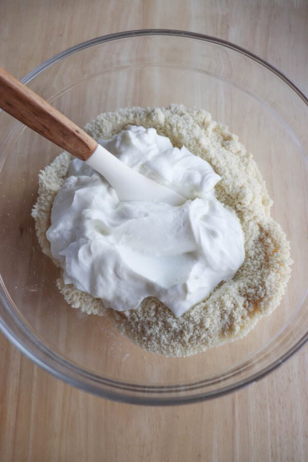 A glass bowl contains a creamy blend of almond flour and yogurt, perfect for crafting gluten-free air fryer bagels. A spatula with a wooden handle rests in the mixture on a wooden surface.