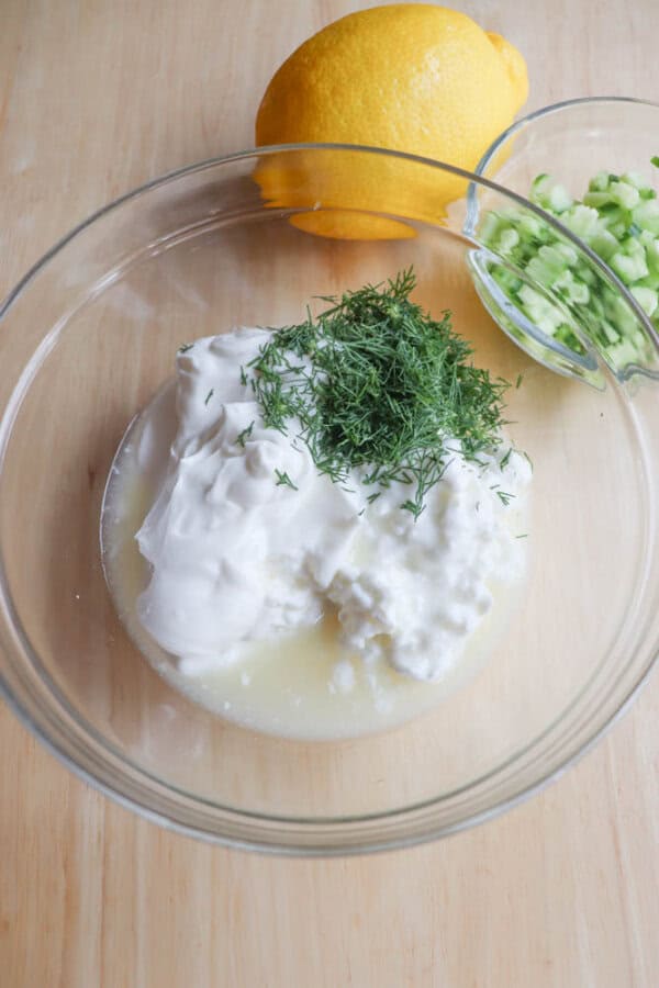 A glass bowl holds a high protein tzatziki mixture of sour cream, cottage cheese, lemon juice, and dill on a wooden surface. Nearby, a whole lemon and chopped cucumbers complete the scene.