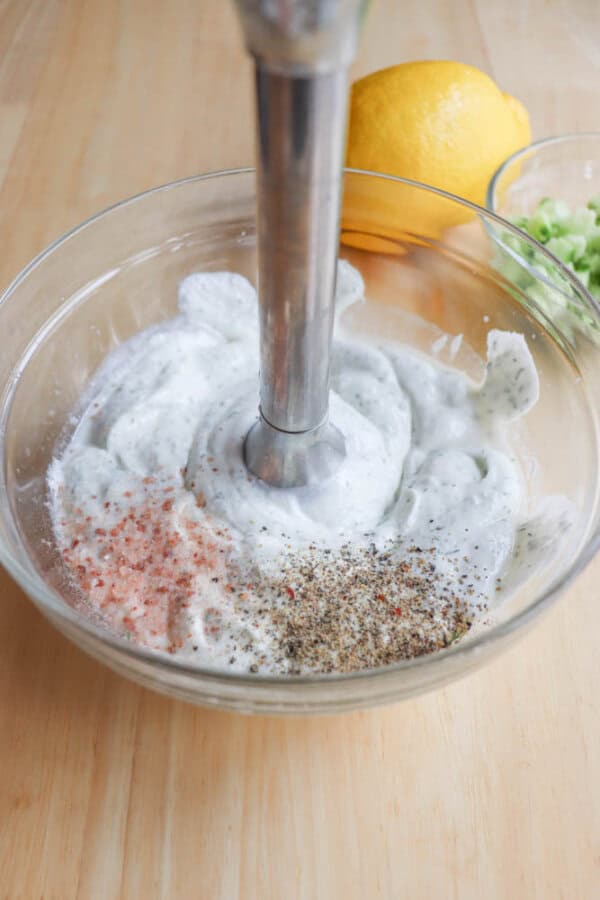 A hand blender mixes a high protein tzatziki recipe, combining yogurt with pink salt, black pepper, and herbs in a glass bowl on a wooden surface. A lemon and chopped green herbs sit invitingly in the background.