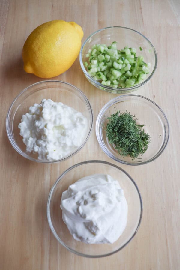 On a wooden surface, the high-protein tzatziki recipe ingredients are laid out: a whole lemon, diced cucumber in a bowl, cottage cheese in another, fresh dill ready to sprinkle, and whipped cream or yogurt to blend it all together.