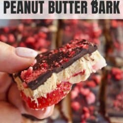 Close-up of a hand holding a slice of strawberry peanut butter bark. The slice has layers of chocolate, peanut butter, and strawberries. More bark pieces are blurred in the background. Text reads Strawberry Peanut Butter Bark and homemadeheather.com.