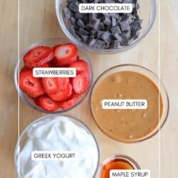 Bowls of ingredients on a wooden surface: dark chocolate chunks, sliced strawberries, creamy peanut butter, Greek yogurt, and a small portion of maple syrup in a container.