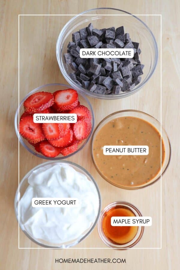 Bowls of ingredients on a wooden surface: dark chocolate chunks, sliced strawberries, creamy peanut butter, Greek yogurt, and a small portion of maple syrup in a container.