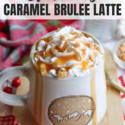 A mug of Starbucks Caramel Brulee Latte topped with whipped cream and caramel drizzle on a wooden surface. Red and white checkered cloth is partially visible. The mug has caramel drips on the sides and sits next to a decorative sprig with red berries.