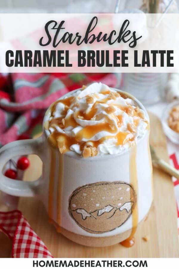 A mug of Starbucks Caramel Brulee Latte topped with whipped cream and caramel drizzle on a wooden surface. Red and white checkered cloth is partially visible. The mug has caramel drips on the sides and sits next to a decorative sprig with red berries.