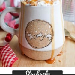 A mug filled with caramel brulee latte topped with whipped cream and caramel drizzle sits on a wooden surface. The mug has a circular design resembling mountains. A checkered cloth, berries, and a syrup bottle are in the background.