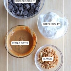 Bowls on a wooden surface contain labeled ingredients: dark chocolate chunks, Greek yogurt, creamy peanut butter, and chopped peanuts. The labels identify each ingredient clearly on the respective bowls.