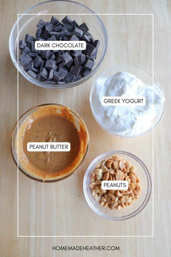 Bowls on a wooden surface contain labeled ingredients: dark chocolate chunks, Greek yogurt, creamy peanut butter, and chopped peanuts. The labels identify each ingredient clearly on the respective bowls.