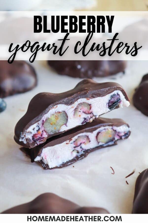 Close-up of two chocolate-covered blueberry yogurt clusters on a parchment paper. One cluster is cut open, revealing blueberries inside creamy yogurt. Text reads Blueberry yogurt clusters at the top and homemadeheather.com at the bottom.